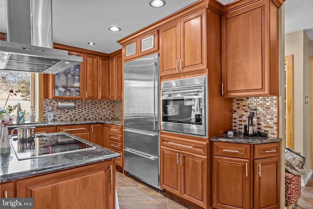 kitchen featuring island exhaust hood, appliances with stainless steel finishes, brown cabinetry, glass insert cabinets, and dark stone counters