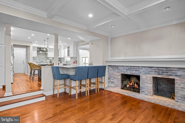 kitchen with beam ceiling, a fireplace, light wood-style flooring, white cabinets, and wall chimney range hood