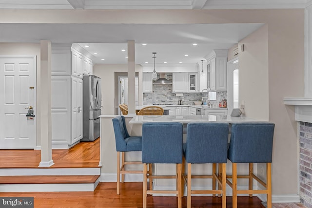 kitchen featuring a kitchen breakfast bar, freestanding refrigerator, a peninsula, wall chimney range hood, and white cabinetry