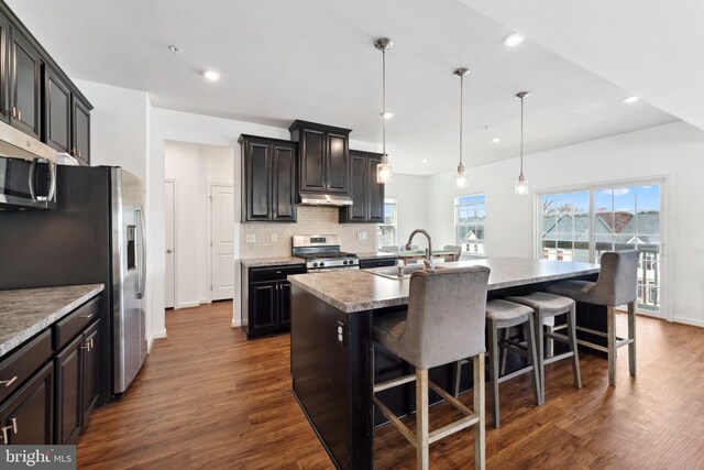 kitchen with decorative backsplash, a kitchen island with sink, appliances with stainless steel finishes, dark wood-type flooring, and a sink