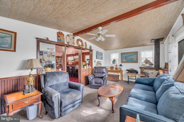 living room featuring lofted ceiling with beams, a wainscoted wall, ceiling fan, a wood stove, and carpet flooring