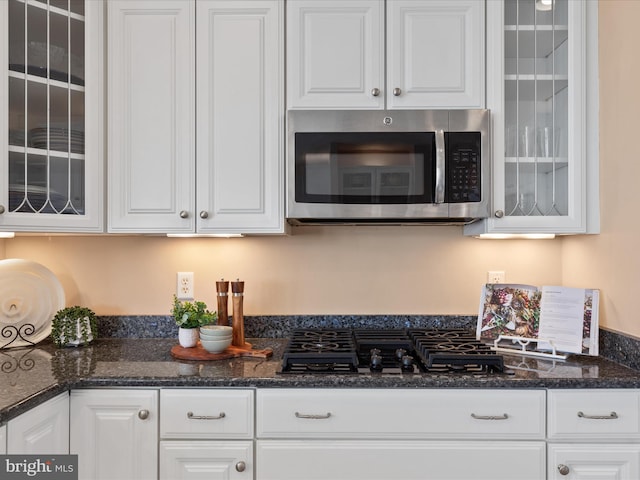 kitchen with black gas cooktop, stainless steel microwave, glass insert cabinets, white cabinetry, and dark stone countertops