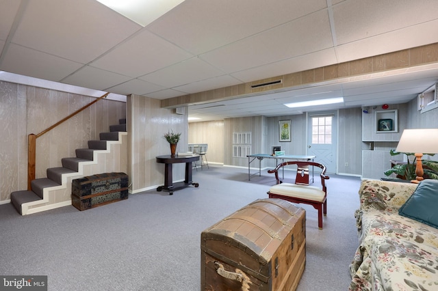 carpeted living area with a paneled ceiling, baseboards, wooden walls, and stairway