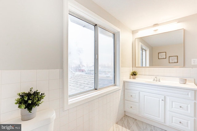 half bathroom with wainscoting, toilet, marble finish floor, vanity, and tile walls