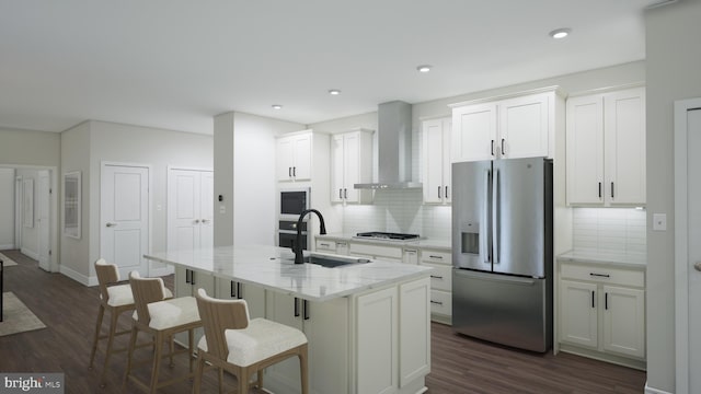 kitchen with appliances with stainless steel finishes, white cabinets, a sink, wall chimney range hood, and an island with sink