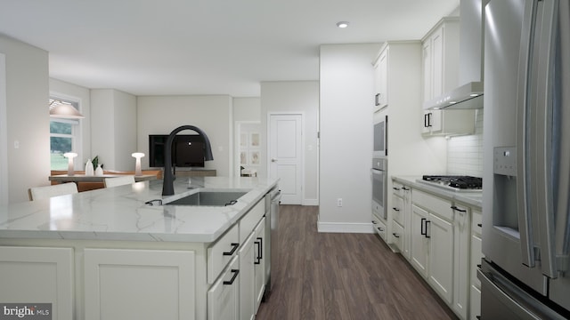 kitchen featuring light stone counters, a kitchen island with sink, stainless steel appliances, a sink, and wall chimney range hood