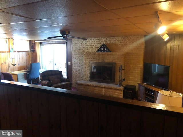 living room featuring a brick fireplace, wooden walls, a ceiling fan, and a drop ceiling