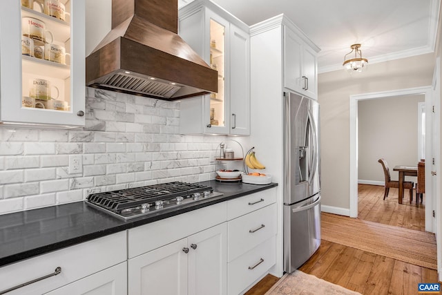 kitchen featuring stainless steel appliances, white cabinetry, wall chimney range hood, dark countertops, and crown molding