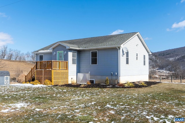 snow covered house with an outbuilding, roof with shingles, a wooden deck, and a storage shed