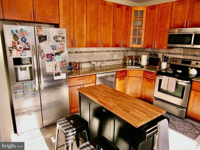 kitchen featuring tasteful backsplash, brown cabinetry, glass insert cabinets, appliances with stainless steel finishes, and dark stone countertops