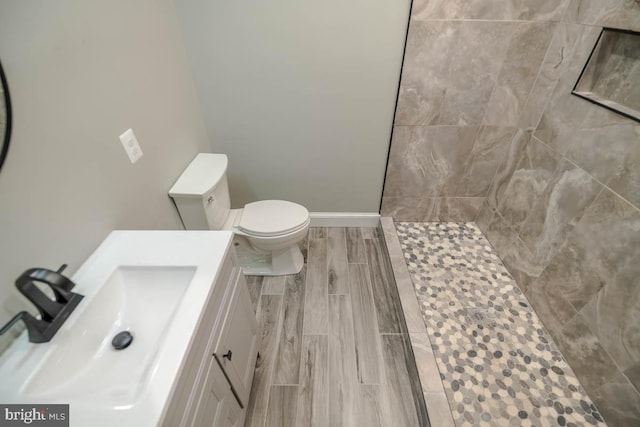 bathroom with baseboards, toilet, wood tiled floor, a tile shower, and vanity