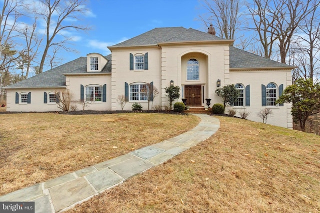 french country home featuring roof with shingles, brick siding, a chimney, and a front yard