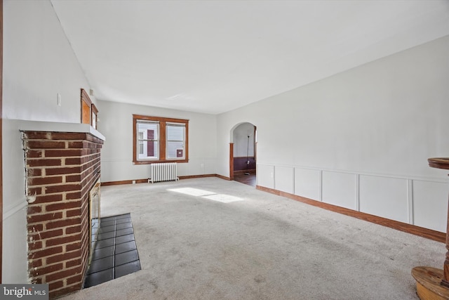 unfurnished living room with arched walkways, a wainscoted wall, radiator, a brick fireplace, and carpet flooring