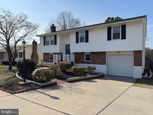 raised ranch with an attached garage, a chimney, concrete driveway, and brick siding