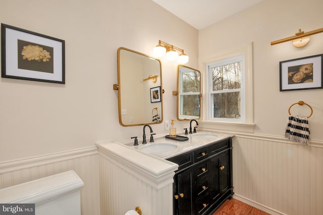 bathroom with a wainscoted wall, a sink, toilet, and double vanity