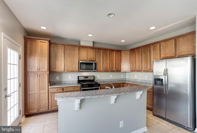 kitchen with appliances with stainless steel finishes, brown cabinets, light stone countertops, an island with sink, and a kitchen bar