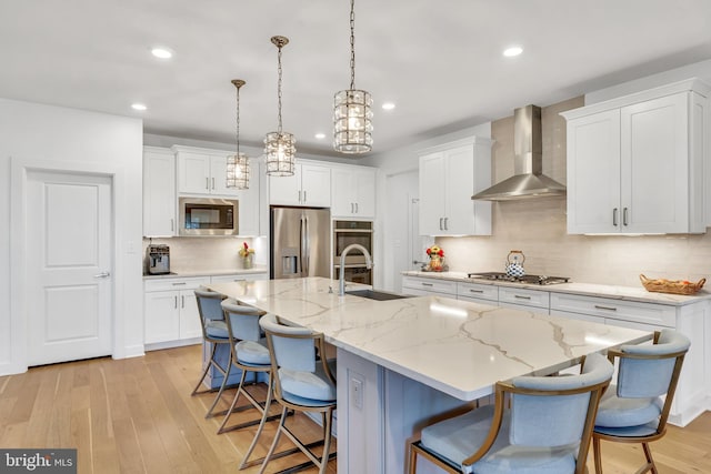 kitchen with stainless steel appliances, hanging light fixtures, a kitchen island with sink, white cabinets, and wall chimney range hood