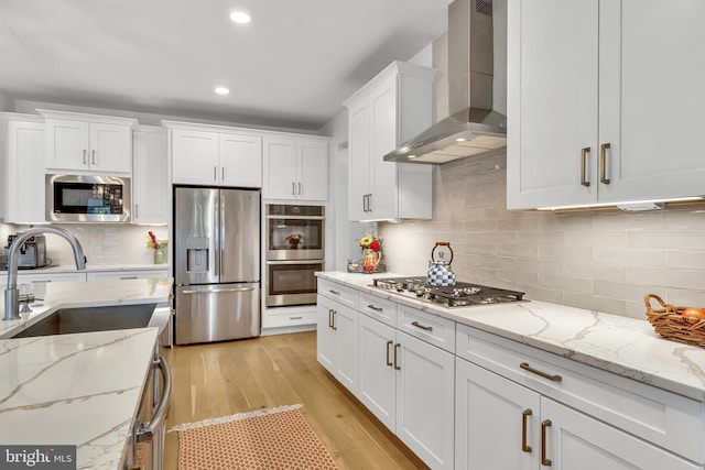 kitchen featuring wall chimney exhaust hood, appliances with stainless steel finishes, light stone countertops, white cabinetry, and a sink