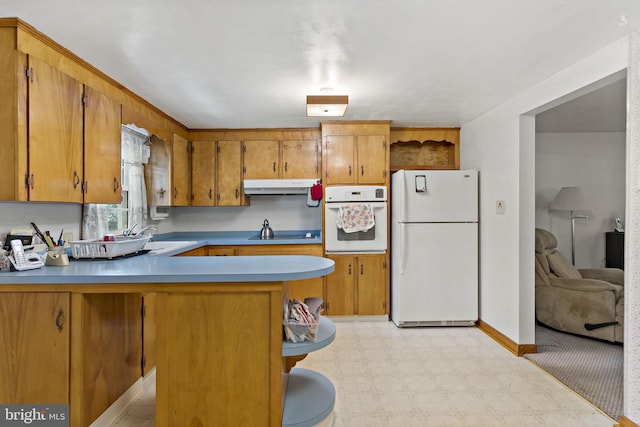 kitchen with under cabinet range hood, light floors, a peninsula, brown cabinetry, and white appliances