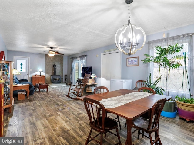 dining room with ceiling fan with notable chandelier, a wood stove, a textured ceiling, and wood finished floors