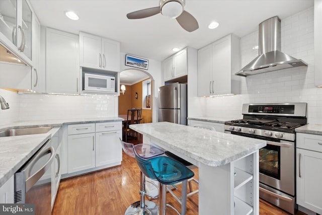 kitchen with arched walkways, wall chimney exhaust hood, appliances with stainless steel finishes, light wood-type flooring, and white cabinetry