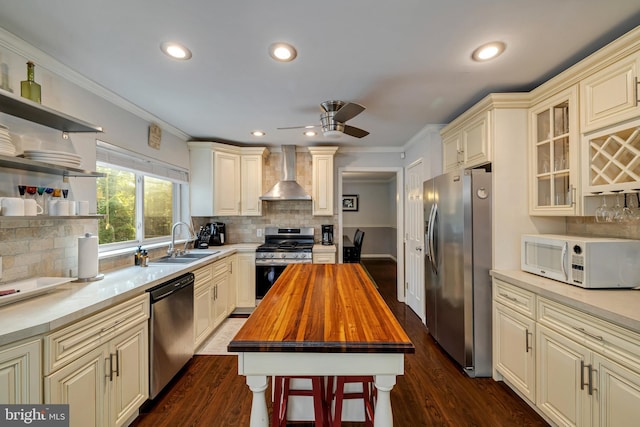 kitchen with stainless steel appliances, butcher block countertops, a sink, cream cabinetry, and wall chimney exhaust hood