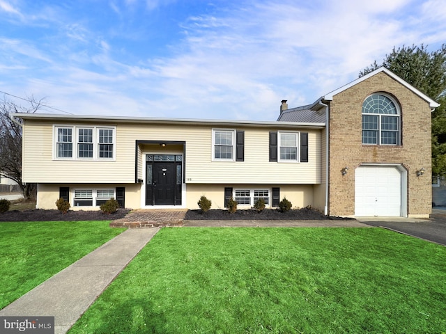 raised ranch featuring a chimney, aphalt driveway, an attached garage, a front lawn, and brick siding