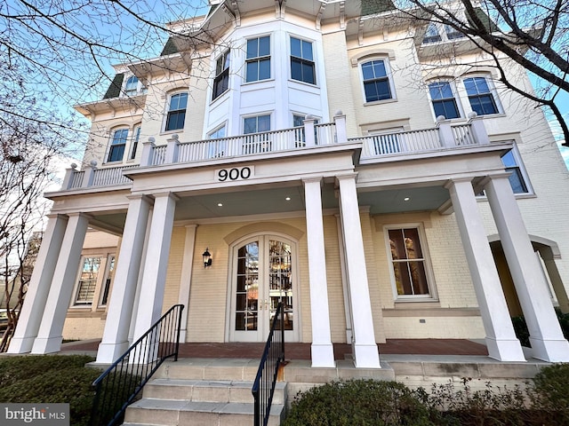 view of exterior entry with covered porch, brick siding, a balcony, and french doors