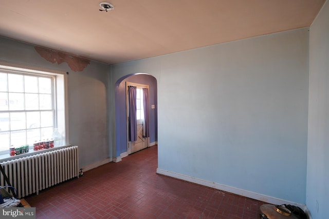 empty room featuring arched walkways, radiator heating unit, brick floor, and baseboards