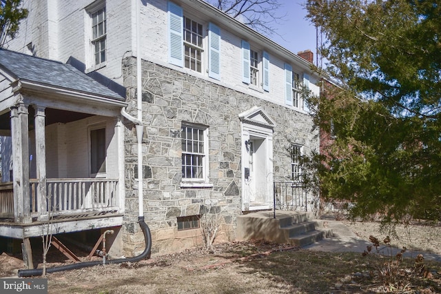 exterior space with a shingled roof, stone siding, covered porch, and a chimney