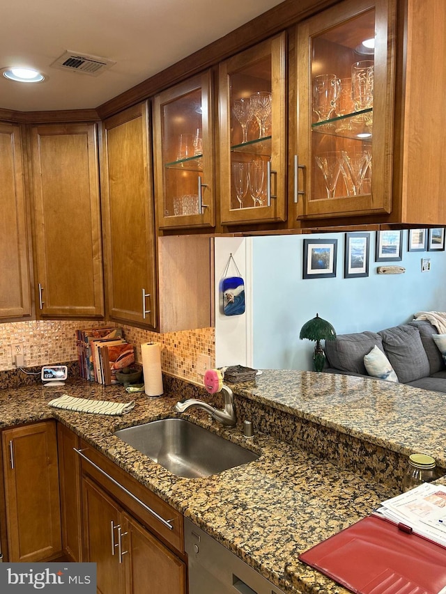 kitchen featuring brown cabinets, decorative backsplash, glass insert cabinets, a sink, and dark stone counters