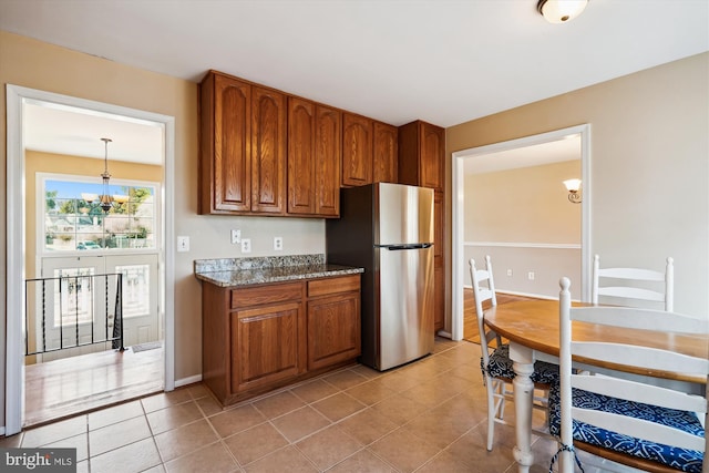 kitchen featuring light tile patterned floors, stone counters, brown cabinets, freestanding refrigerator, and an inviting chandelier