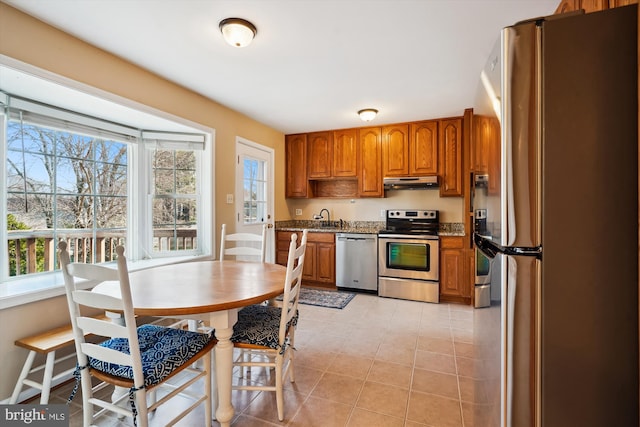 kitchen with under cabinet range hood, a sink, appliances with stainless steel finishes, light tile patterned flooring, and brown cabinetry