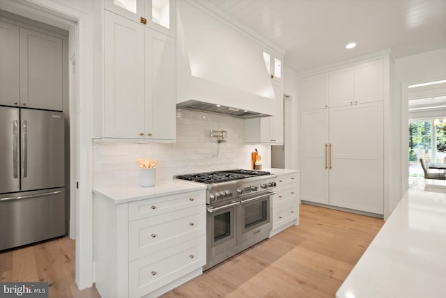 kitchen featuring premium range hood, appliances with stainless steel finishes, light wood finished floors, glass insert cabinets, and tasteful backsplash