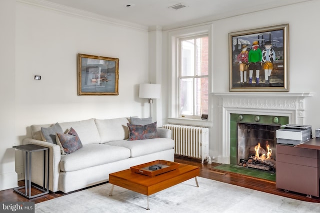 living room featuring visible vents, radiator, crown molding, a warm lit fireplace, and wood finished floors