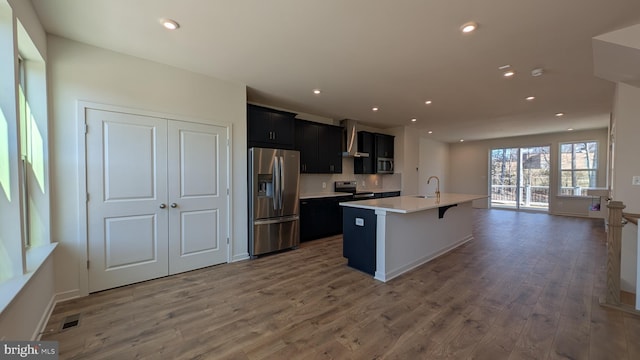 kitchen featuring a kitchen island with sink, stainless steel appliances, wood finished floors, a sink, and dark cabinetry