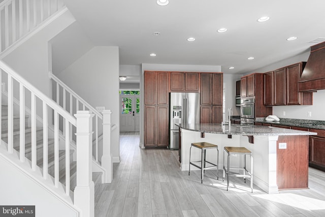 kitchen featuring stainless steel appliances, light wood-type flooring, premium range hood, and an island with sink