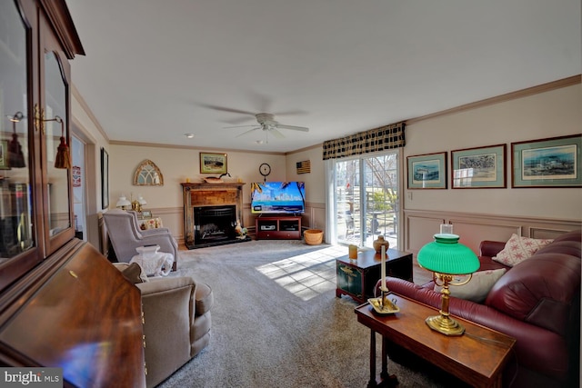 carpeted living room featuring a wainscoted wall, ceiling fan, crown molding, a fireplace, and a decorative wall