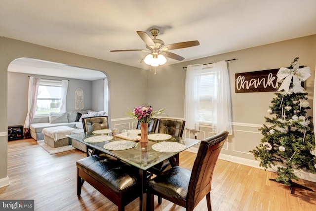 dining space with arched walkways, ceiling fan, a decorative wall, wood finished floors, and wainscoting