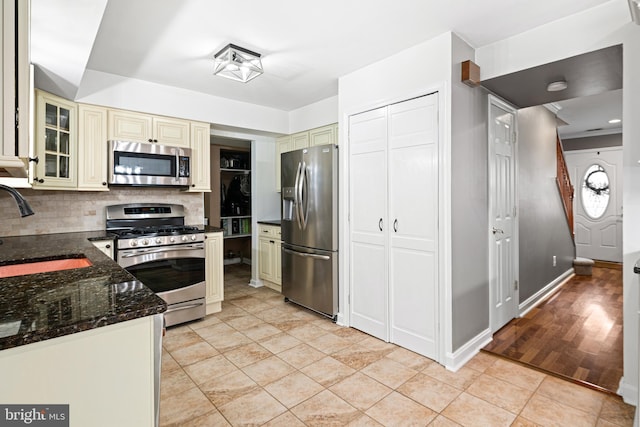 kitchen with cream cabinetry, a sink, stainless steel appliances, dark stone counters, and decorative backsplash