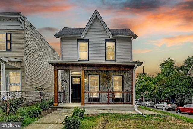 view of front of home featuring stone siding, covered porch, roof with shingles, and fence