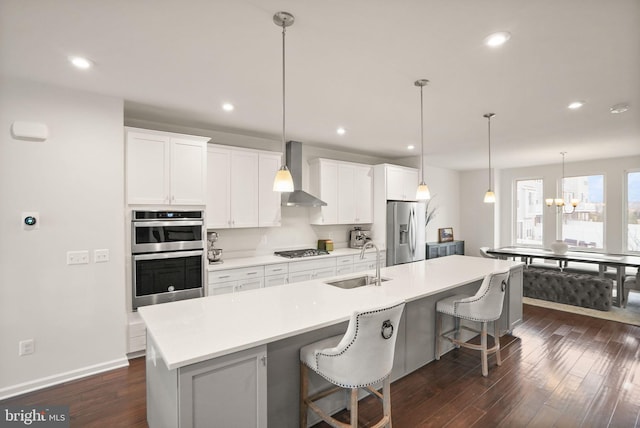 kitchen featuring dark wood finished floors, an island with sink, a sink, stainless steel appliances, and wall chimney exhaust hood