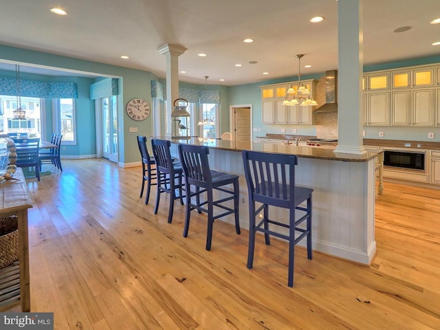 kitchen featuring light wood finished floors, wall chimney exhaust hood, cream cabinets, ornate columns, and black microwave