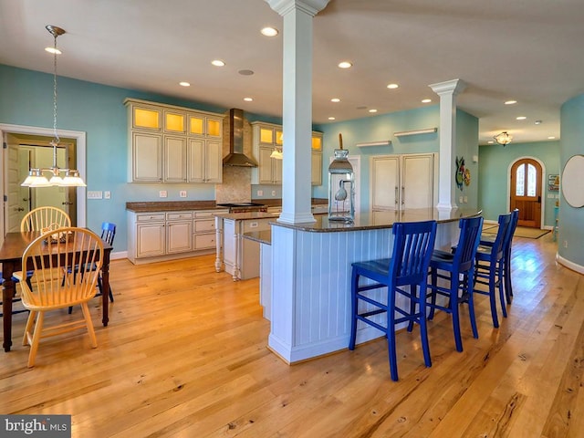 kitchen featuring light wood-type flooring, decorative columns, wall chimney range hood, and a breakfast bar area