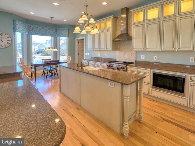 kitchen with a center island with sink, wall chimney exhaust hood, cream cabinetry, light wood-type flooring, and a sink