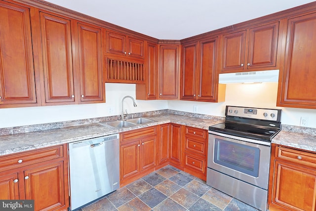 kitchen featuring under cabinet range hood, stainless steel appliances, a sink, brown cabinetry, and stone finish floor