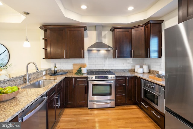 kitchen featuring a tray ceiling, light wood finished floors, stainless steel appliances, a sink, and wall chimney range hood