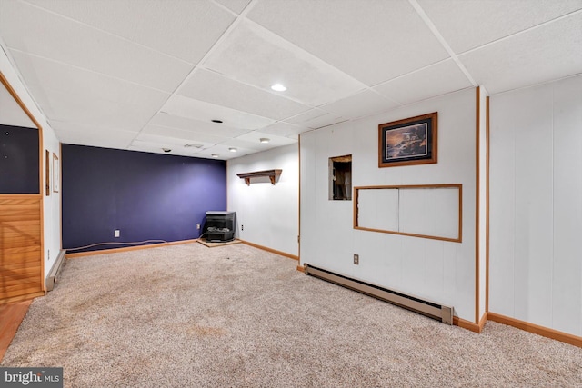 below grade area with a baseboard heating unit, a drop ceiling, carpet, and a wood stove