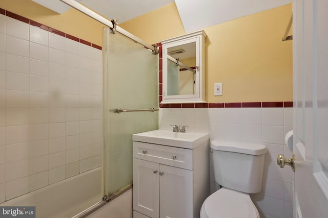 bathroom featuring toilet, a wainscoted wall, combined bath / shower with glass door, vanity, and tile walls