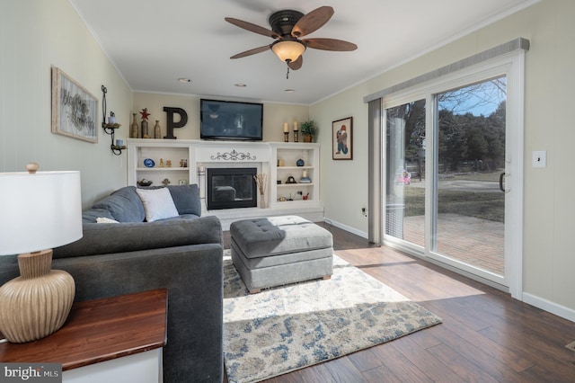 living area featuring baseboards, a glass covered fireplace, ceiling fan, ornamental molding, and wood finished floors